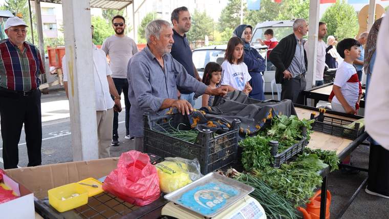 Köy pazarında tezgahın önünde bir adam, sebze ve yeşilliklerle dolu kasaların arasında duruyor.