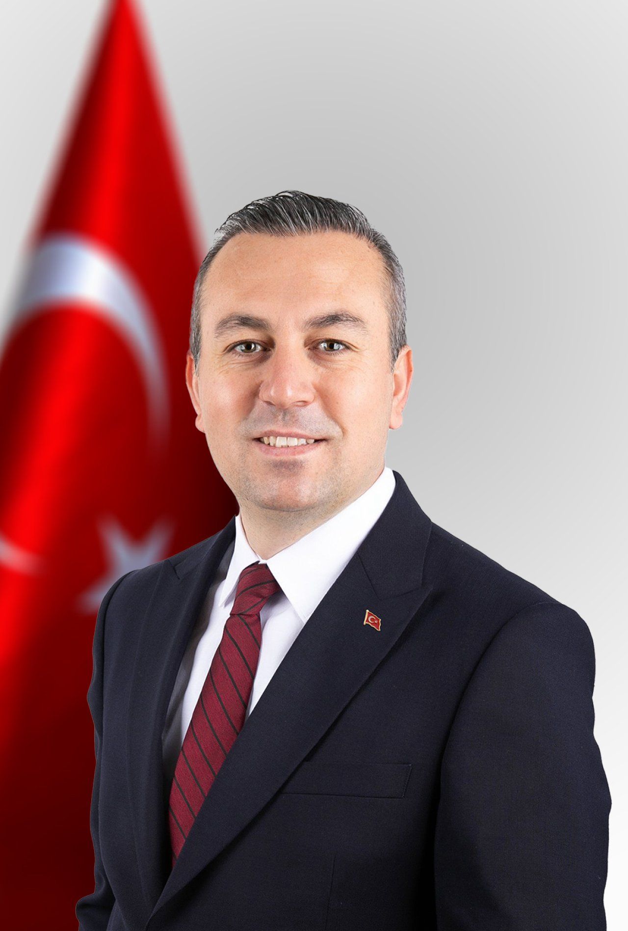 Takım elbiseli bir erkek, arkasında Türk bayrağı bulunan stüdyo portresinde kameraya bakıyor.