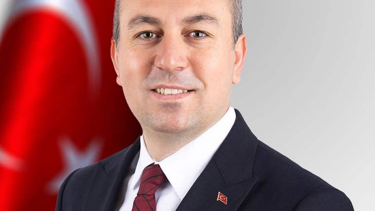Takım elbiseli bir erkek, arkasında Türk bayrağı bulunan stüdyo portresinde kameraya bakıyor.