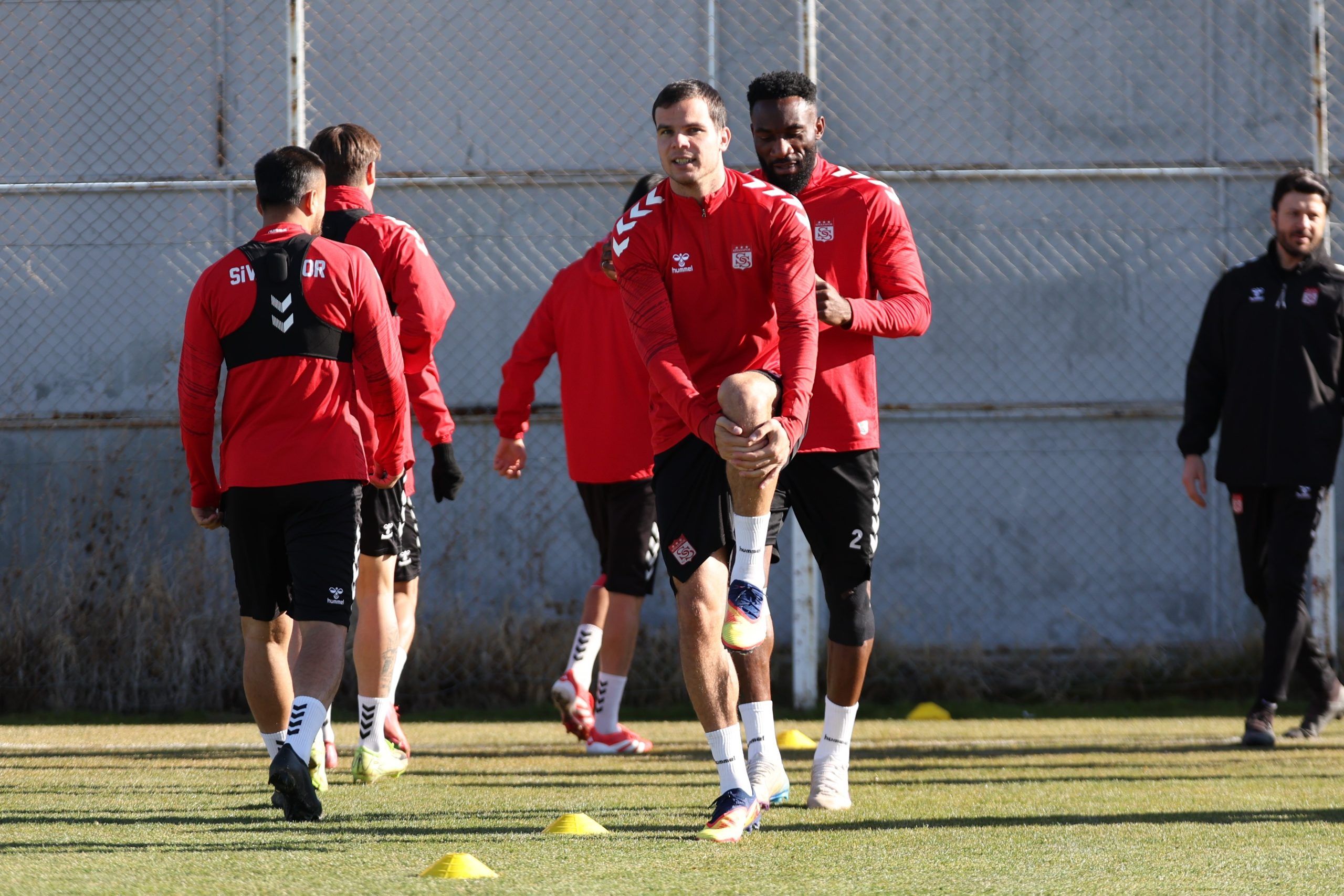 Kırmızı forma giymiş futbolcular, antrenman sahasında ısınma hareketleri yapıyor.
