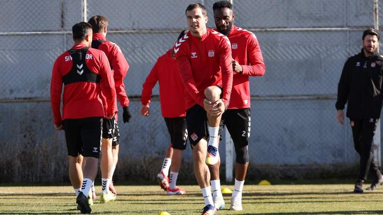 Kırmızı forma giymiş futbolcular, antrenman sahasında ısınma hareketleri yapıyor.