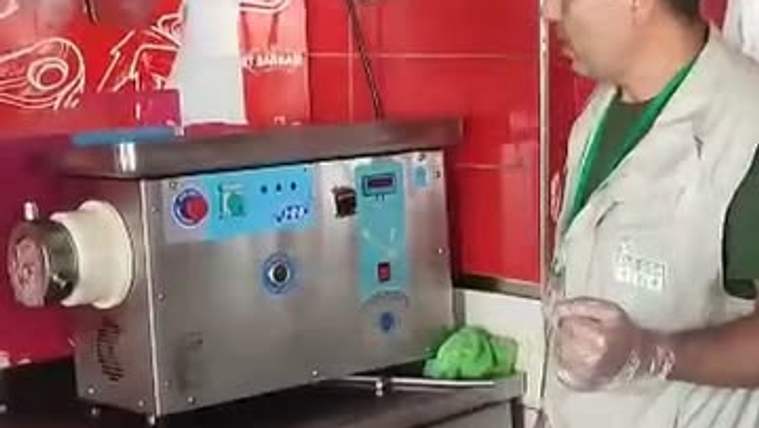 Kırmızı duvarlı bir alanda, eldivenli bir kişi et kıyma makinesinin yanında duruyor; tezgâhta yeşil poşet bulunuyor.