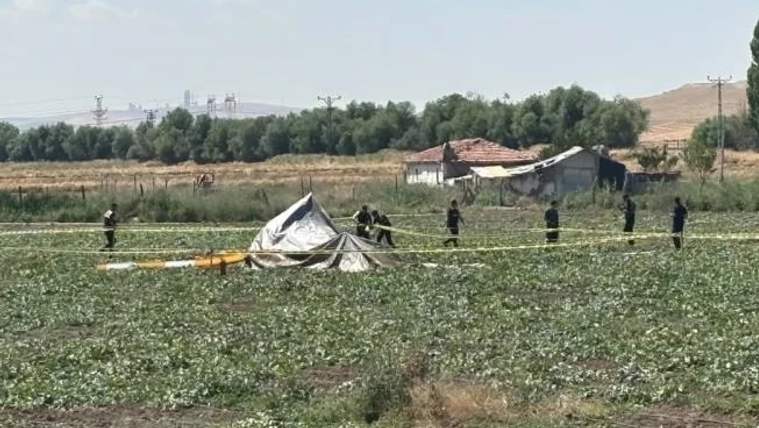 Ekili bir tarlada devrilmiş beyaz bir araç parçası, güvenlik şeridi ve çevresinde duran kişiler görülüyor.
