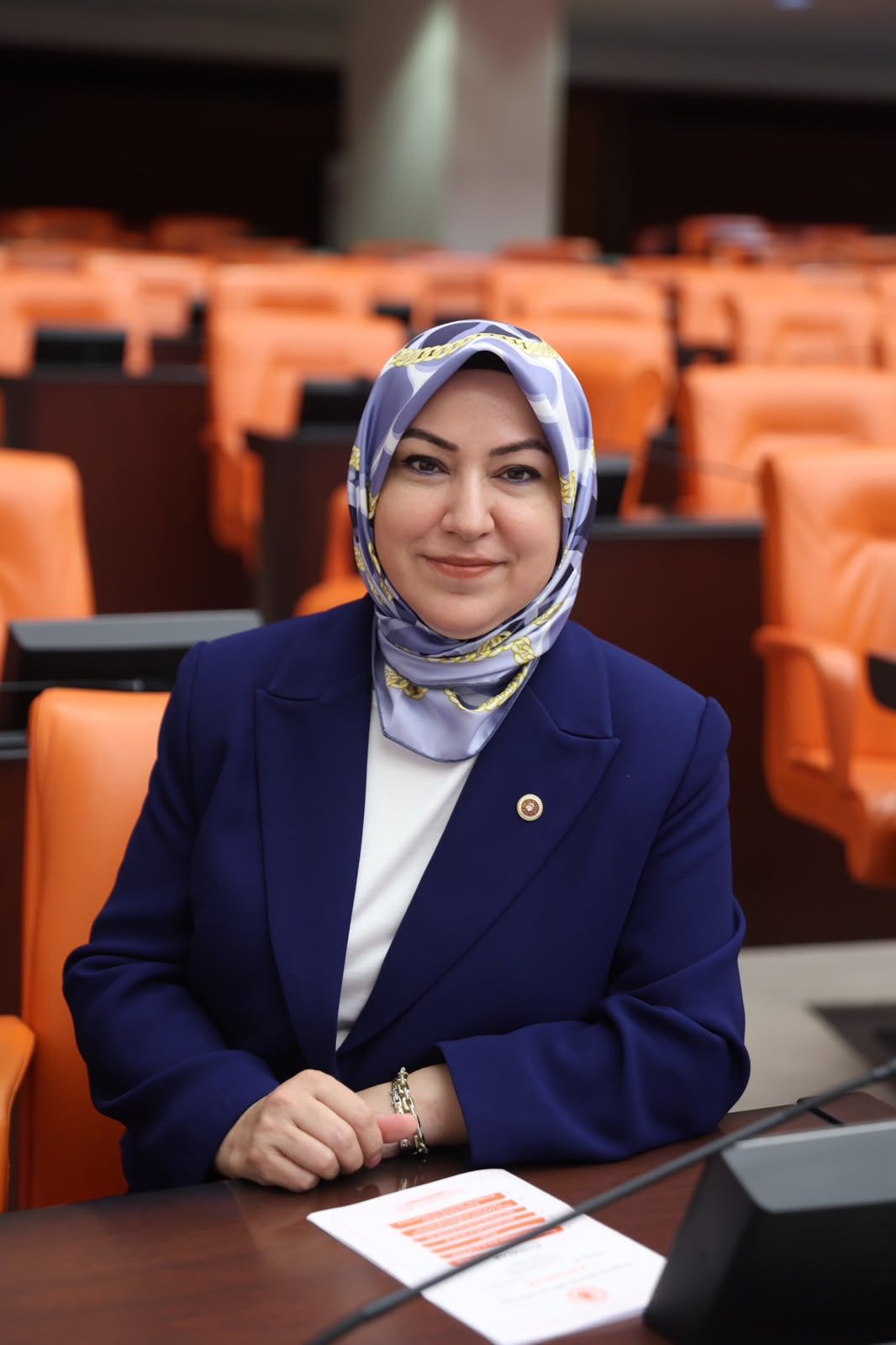 Mavi bir başörtüsü takan bir kadın, parlamento koltuklarının önünde gülümsüyor.