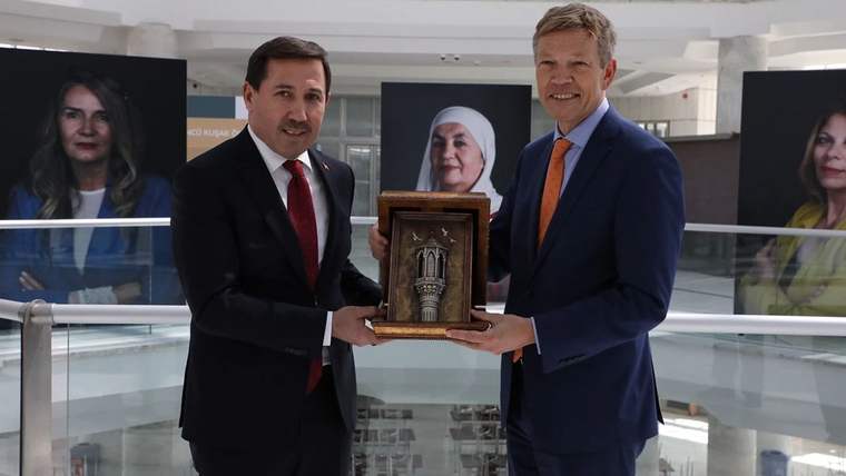 İki erkek, sergi alanında üzerinde kabartmalı motif bulunan ahşap bir plaketi birlikte tutuyor; arkada portreler görülüyor.