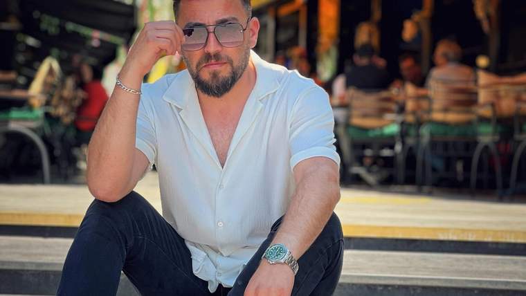 Siyah pantolon ve beyaz gömlek giymiş bir erkek, otururken güneş gözlüğü takıyor.
