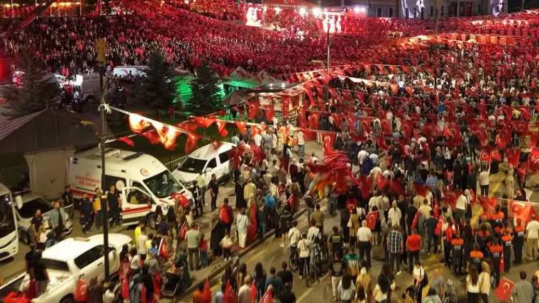 Kalabalık, Türk bayraklarıyla süslenmiş sokakta toplanmış. İtfaiye aracı ve ambulans görünmekte.