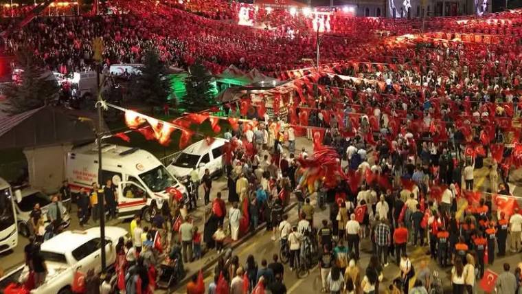 Kalabalık, Türk bayraklarıyla süslenmiş sokakta toplanmış. İtfaiye aracı ve ambulans görünmekte.