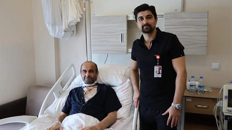 Hastane odasında, hasta yatakta otururken yanında bir sağlık çalışanı duruyor.