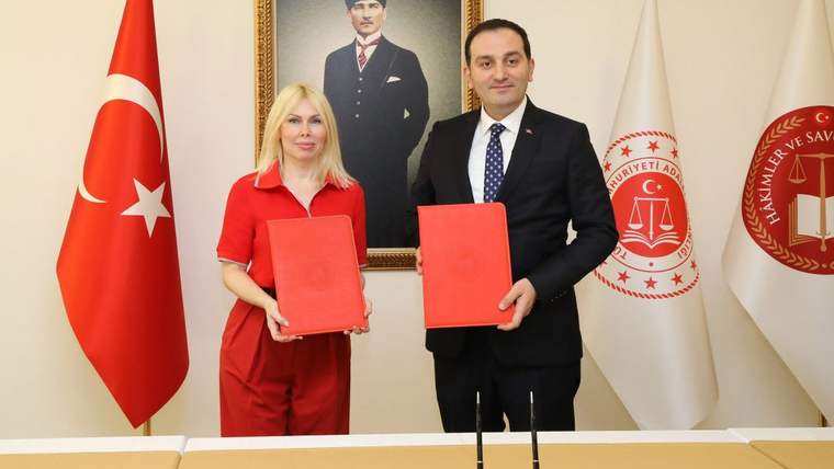 İki kişi, Türk bayrağı ve adalet amblemli bayrakların önünde kırmızı dosyalar tutuyor.