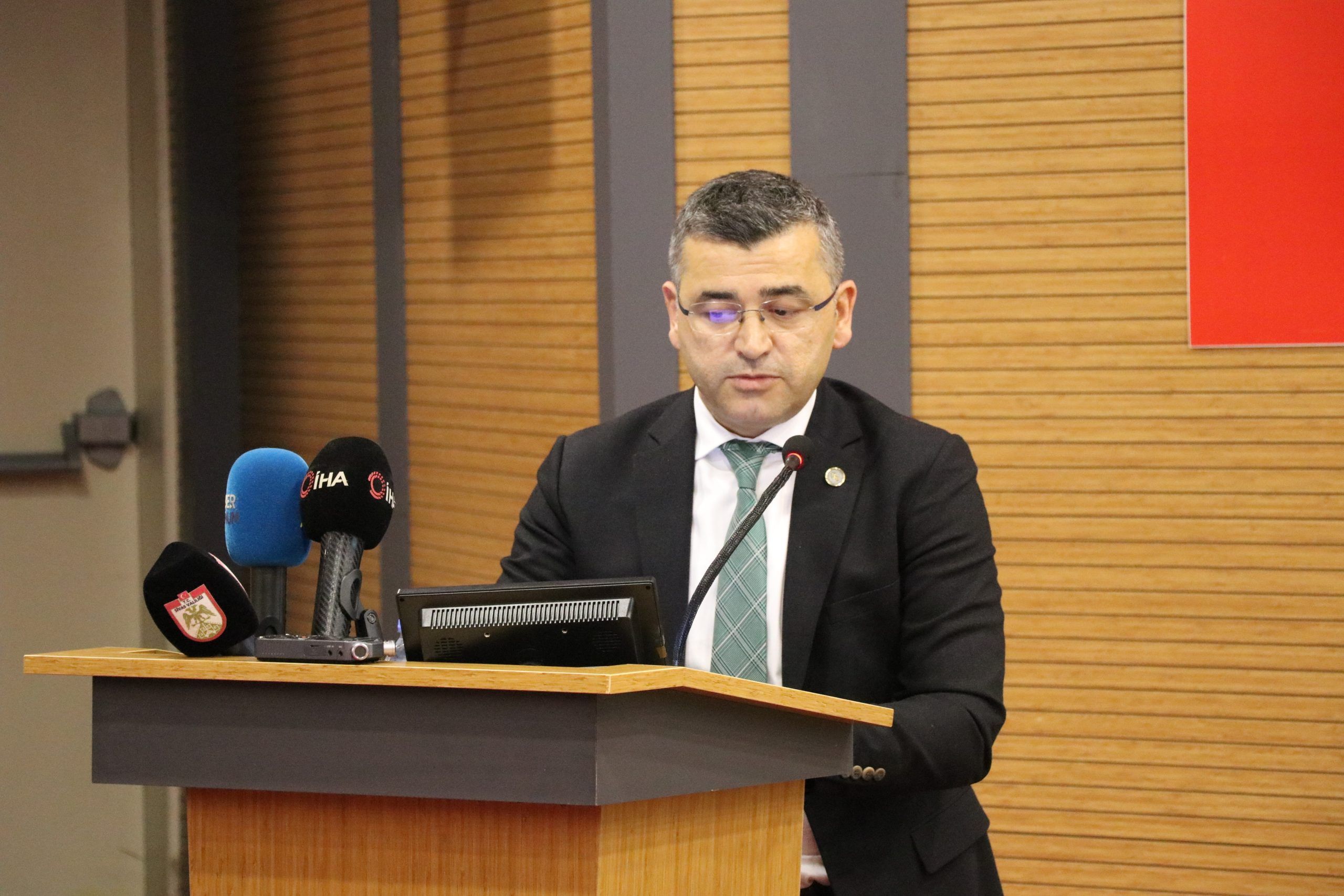 Kürsüde konuşan bir kişi, önünde mikrofon ve mavi renkli mikrofonlar var.