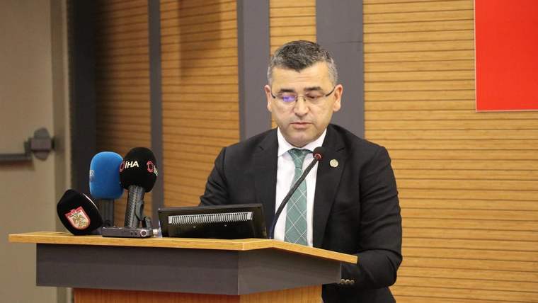 Kürsüde konuşan bir kişi, önünde mikrofon ve mavi renkli mikrofonlar var.