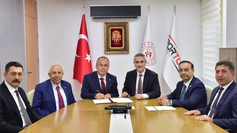 Bir masanın etrafında oturan altı kişi, belgeleri imzalıyor. Arkada Türk bayrağı ve T.C. arması var.