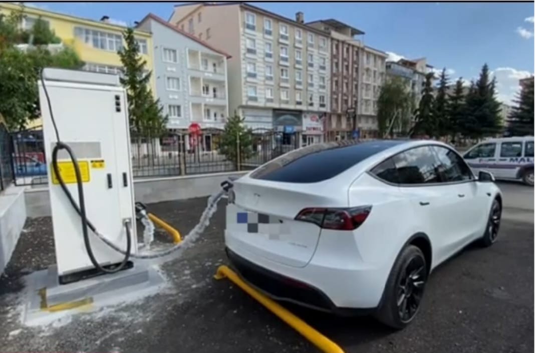 Beyaz bir SUV, elektrikli araç şarj ünitesinin yanında; arka planda çok katlı binalar görülüyor.