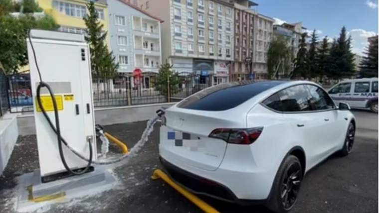 Beyaz bir SUV, elektrikli araç şarj ünitesinin yanında; arka planda çok katlı binalar görülüyor.