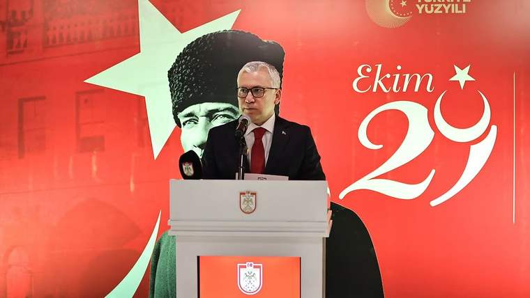 Gözlüklü bir erkek, üzerinde arma bulunan kürsüde konuşuyor; arka planda Atatürk portresi ve “Ekim 29” yazısı görülüyor.
