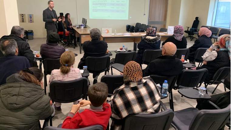 Toplantı salonunda bir kişi sunum yaparken, karşısında oturan katılımcılar projeksiyon ekranını izliyor.