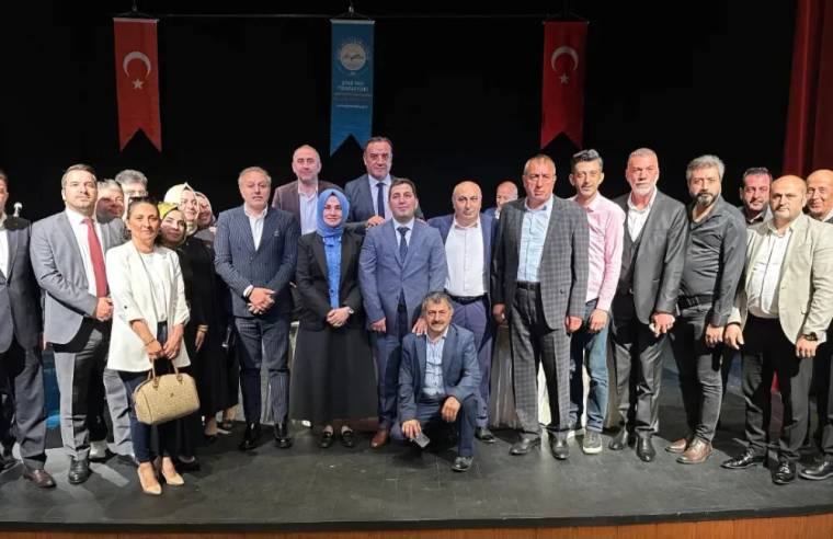 Sahne üzerinde Türk bayrakları ve flamalar önünde poz veren bir grup insan, önde oturan bir kişiyle birlikte görülüyor.
