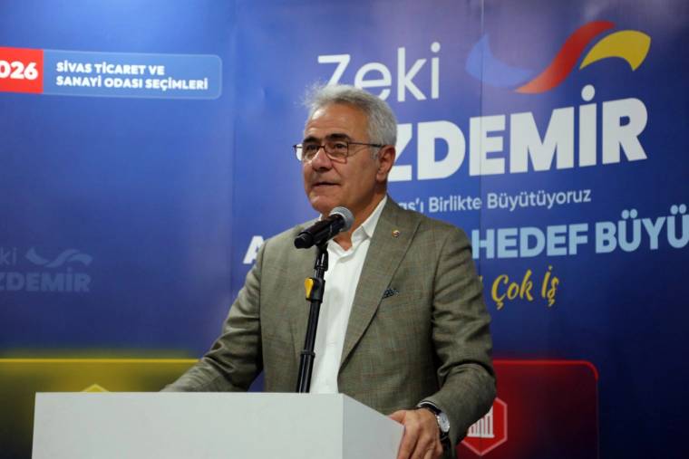 Kürsüde konuşan gözlüklü bir erkek, üzerinde yazılar bulunan mavi bir fonun önünde duruyor.