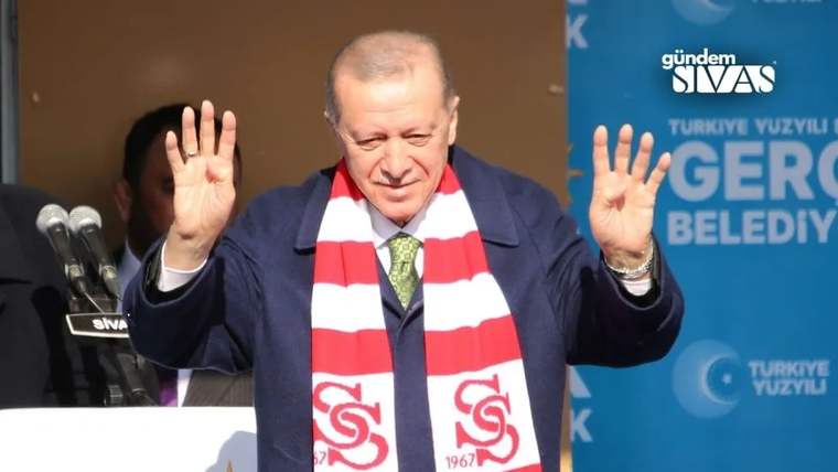 Kırmızı-beyaz atkılı bir kişi, kürsü önünde iki elini kaldırırken arka planda mavi pankart bulunuyor.