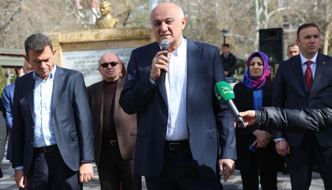 Açık alanda elinde mikrofonla konuşan bir kişi, çevresinde duran kadın ve erkeklerle birlikte görülüyor.