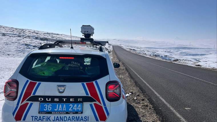 Karlı yol kenarında duran, üzerinde “Trafik Jandarması” yazılı araç ve tavana monte radar cihazı görülüyor.