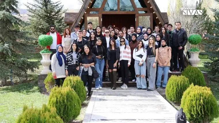Bir grup genç, bir ahşap yapının önünde poz veriyor; arka planda ağaçlar ve çalılar var.