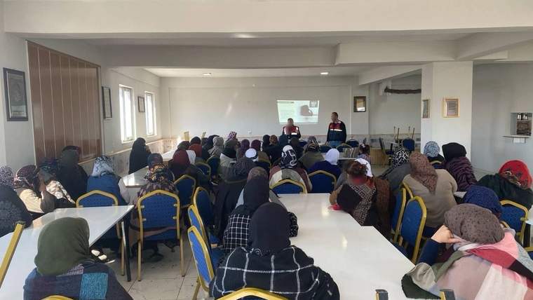 Toplantı salonunda çok sayıda katılımcı sunumu izlerken önde iki kişi kürsü yanında duruyor.