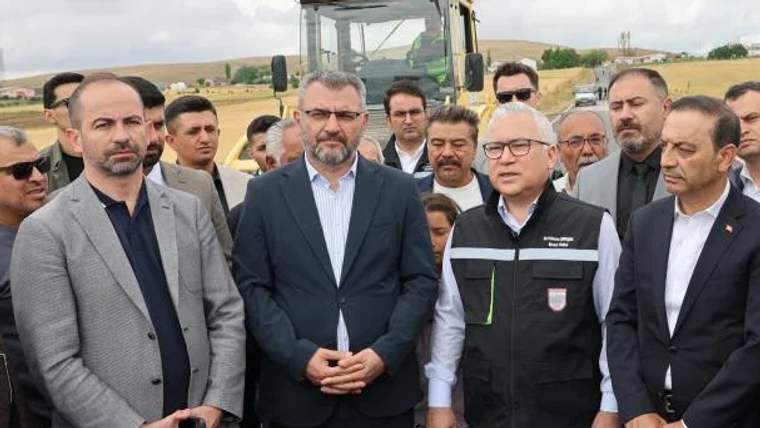 Bir grup insan, asfaltlama çalışmaları öncesinde bir araya gelmiş. Arkada asfalt dökme aracı görünüyor.