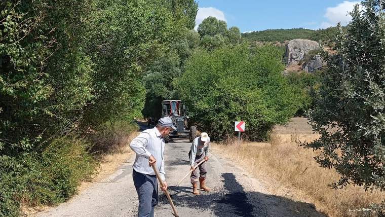 Köy yolunda asfalt çalışması yapan iki kişi, bir iş makinesi arka planda görünüyor.