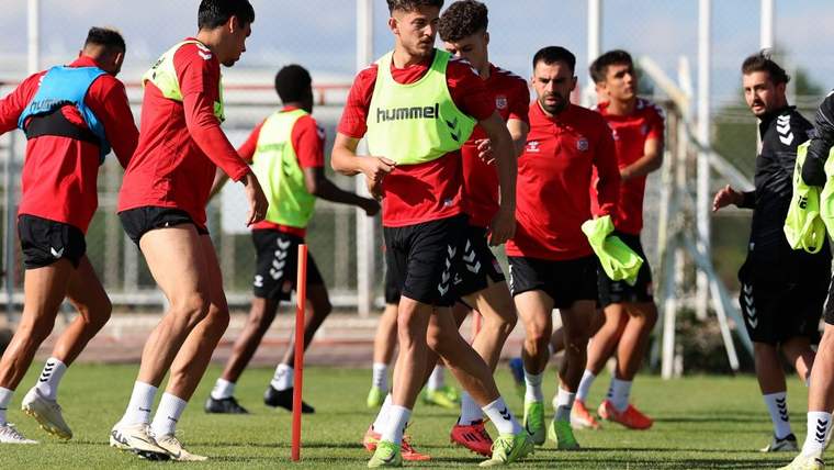 Kırmızı antrenman kıyafetleri ve yeşil yelekler giyen futbolcular, sahada antrenman yapıyor.