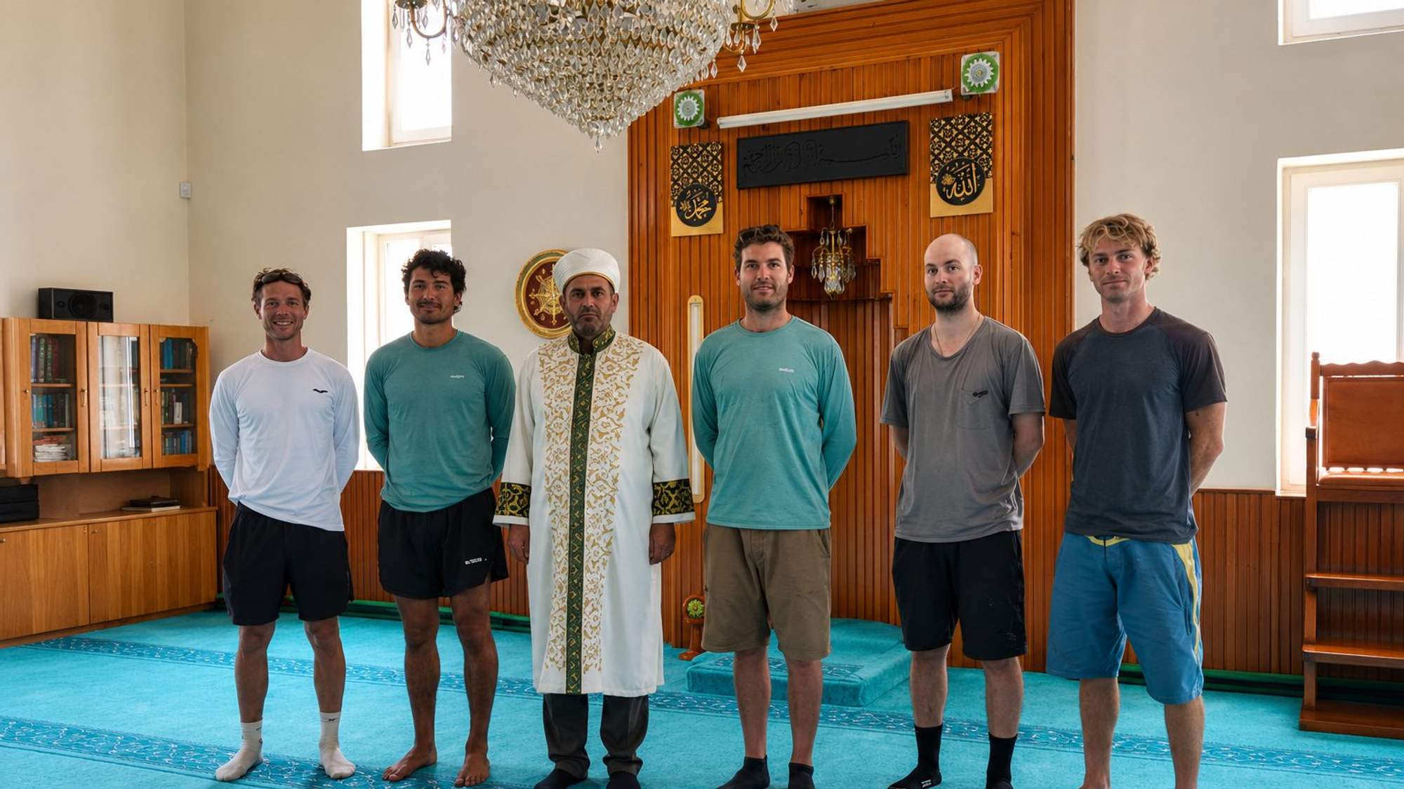 Cami içinde, işlemeli cübbeli bir kişi ve beş kişi mihrap önünde yan yana duruyor.