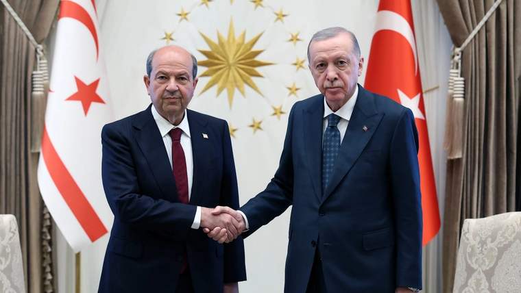 İki erkek, Türk ve KKTC bayrakları önünde takım elbiseleriyle el sıkışıyor.