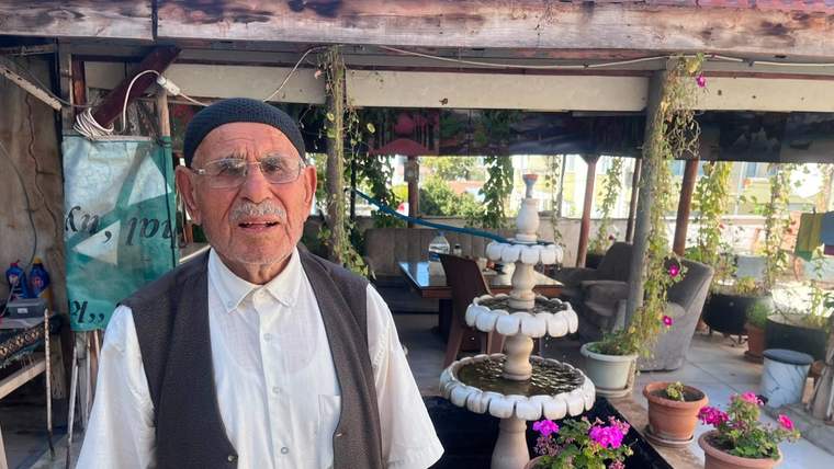 Siyah takkeli ve gözlüklü yaşlı bir adam, çiçek saksıları ve süs havuzunun önünde duruyor.