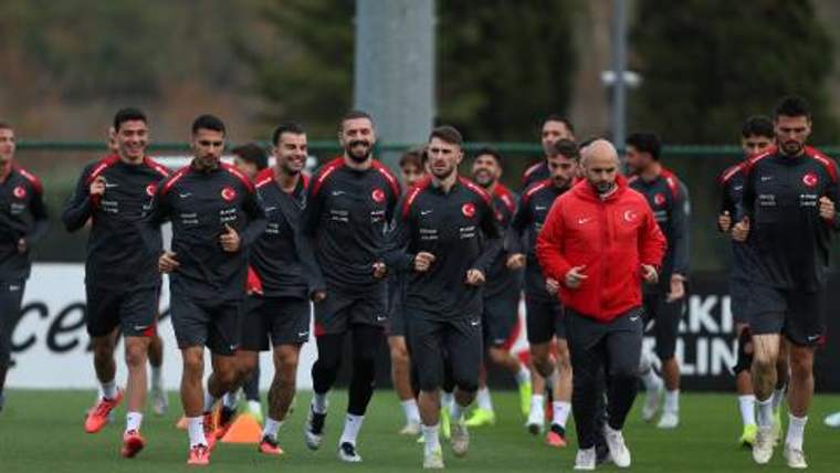Futbolcular, antrenman sahasında siyah formaları ve kırmızı montlu bir kişiyle birlikte koşuyor.