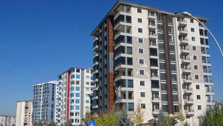 Mavi gökyüzü altında, balkonlu ve çok katlı birkaç apartman binası yan yana görülüyor.