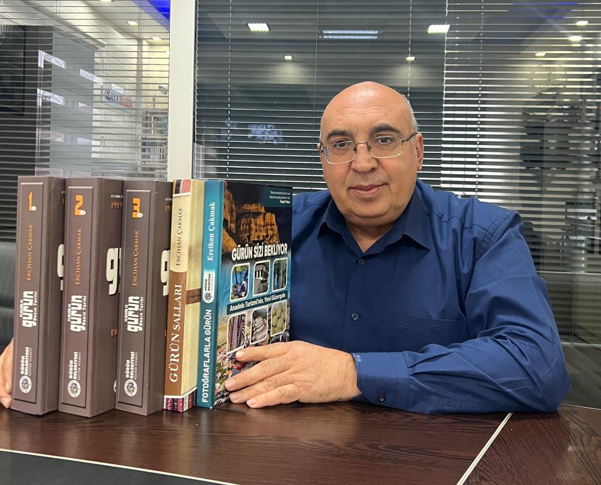 Bir kişi, elinde kitaplarla birlikte bir masada oturuyor. Arkada camlı bir ofis görünmekte.