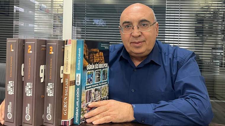Bir kişi, elinde kitaplarla birlikte bir masada oturuyor. Arkada camlı bir ofis görünmekte.