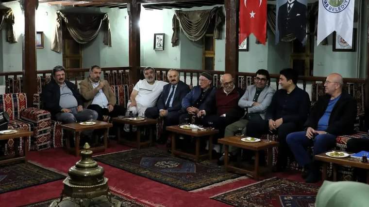 Türk bayrağı ve belediye flamalarının bulunduğu salonda, koltuklarda oturan bir grup kişi görülüyor.