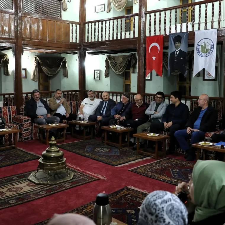 Türk bayrağı ve belediye flamalarının bulunduğu salonda, koltuklarda oturan bir grup kişi görülüyor.