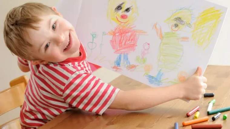 Bir çocuk, elinde çizdiği resimle gülümseyerek poz veriyor. Masanın üzerinde renkli pastel boyalar var.