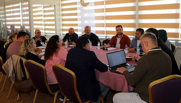 Toplantı masası çevresinde oturan kişiler, önlerindeki belgeler ve açık bir dizüstü bilgisayarla görüşüyor.