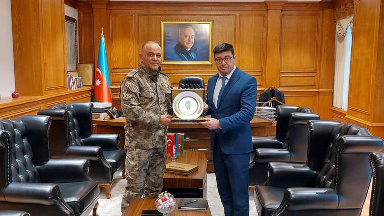 Ahşap panelli bir odada kamuflaj üniformalı ve takım elbiseli iki kişi, plaket tutarak yan yana duruyor.