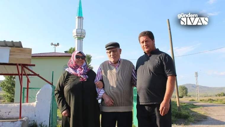 Başörtülü bir kadın, kasketli yaşlı bir erkek ve bir adam, cami minaresi önünde yan yana duruyor.