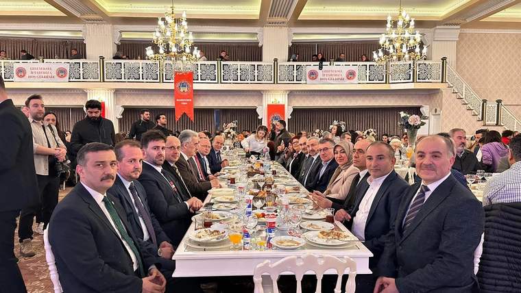 Uzun bir masada oturan kalabalık bir grup, iftar yemeği için hazırlanmış. Masada yiyecek ve içecekler var.