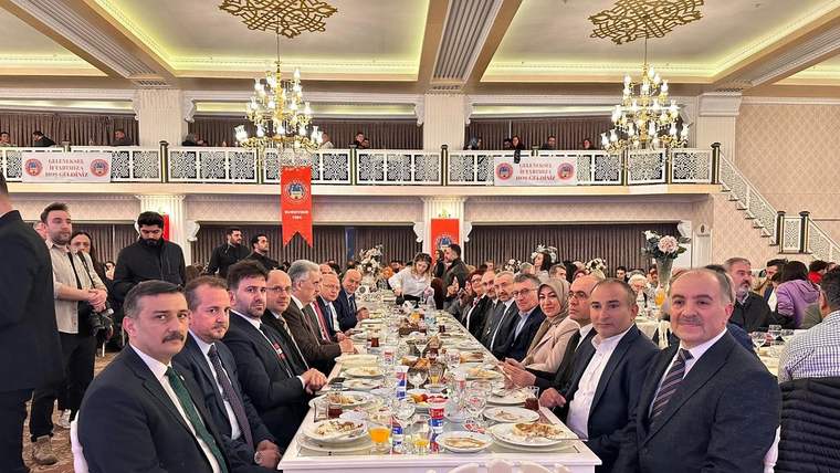 Uzun bir masada oturan kalabalık bir grup, iftar yemeği için hazırlanmış. Masada yiyecek ve içecekler var.