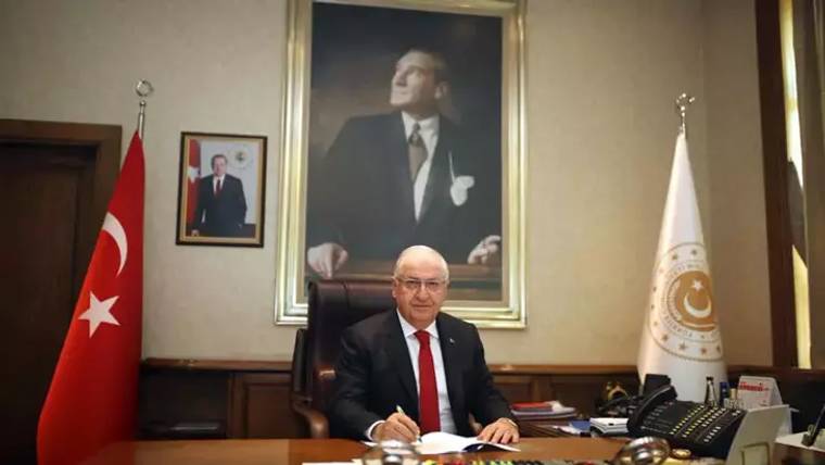 Masa başında oturan bir kişi, arkasında büyük bir portre ve iki bayrak bulunan odada evrak imzalıyor.