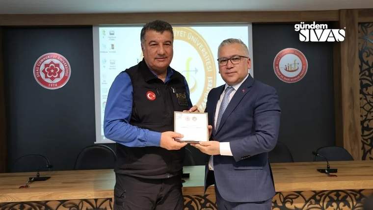 Toplantı salonunda iki erkek, bir belgeyi birlikte tutarken arkalarında projeksiyon ve kurum amblemleri görülüyor.