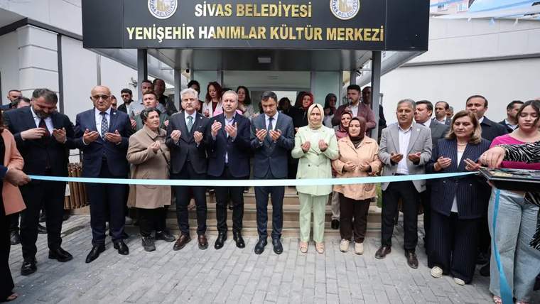 Kurdele kesme töreninde bir grup insan, dua ederken ve kurdeleyi tutarken görüntüleniyor.