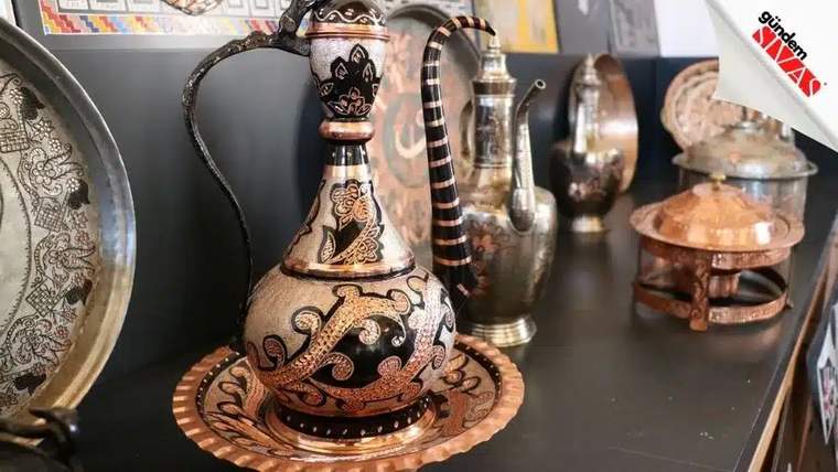 Siyah, bakır ve altın renkli işlemelerle süslü ibrik, işlemeli yuvarlak bir tepsi üzerinde sergileniyor.